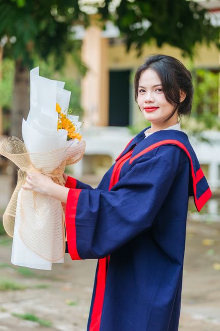 A young woman dressed in a navy blue graduation gown with red accents stands outdoors, holding a floral gift of yellow and white flowers wrapped in white paper and beige mesh, tied with a natural twin
