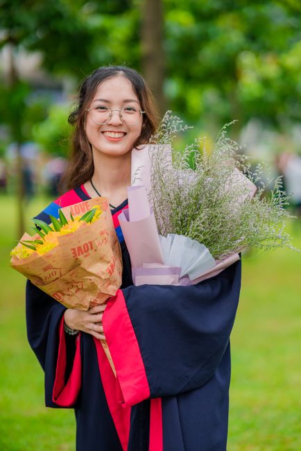 A young woman smiling outdoors in a park during daytime, wearing a graduation gown with red and black accents. She holds two bouquets: one wrapped in colorful paper with yellow, blue, and red elements