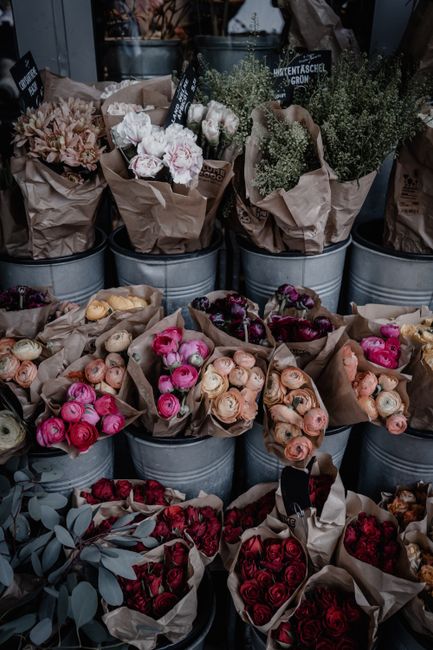 A display of various fresh flowers arranged in galvanized metal buckets and wrapped in brown kraft paper, featuring a range of vibrant colours including soft pinks, creamy whites, deep reds, and purpl