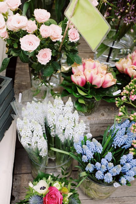 A display of fresh flowers arranged in various vases and containers on a wooden surface, featuring light pink roses with lush green foliage, pink and cream tulips with broad green leaves, clusters of 