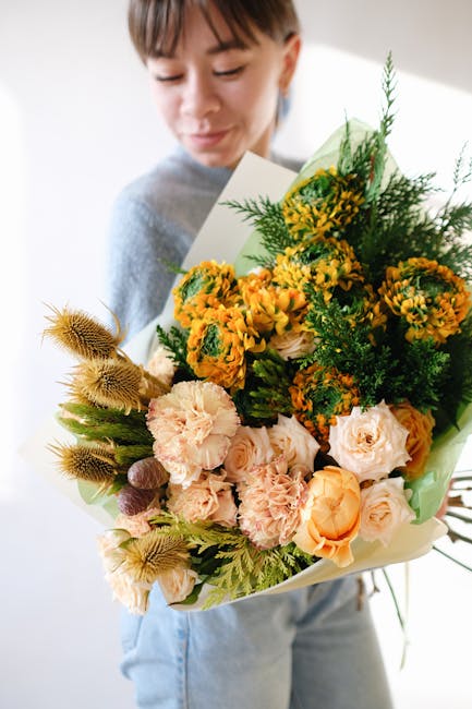 A woman in a light grey sweater holding a large, vibrant floral gift bouquet in her hands. The arrangement features bright yellow and orange flowers, including roses, carnations, and small chrysanthem