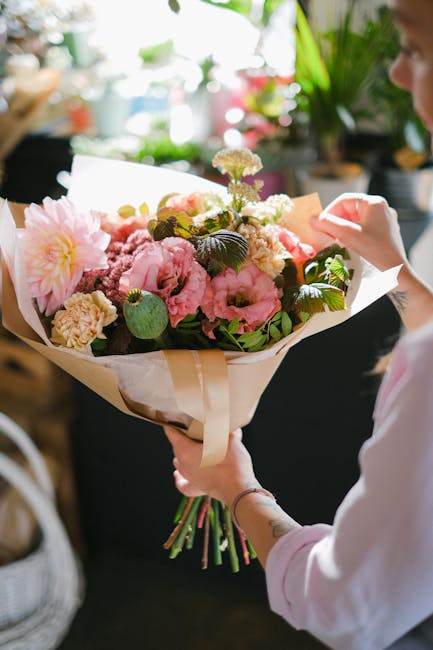 A person holding a vibrant floral bouquet featuring pink peonies, white ranunculus, and greenery wrapped in cream and white tissue paper, with additional floral accents on top. The bouquet is large an