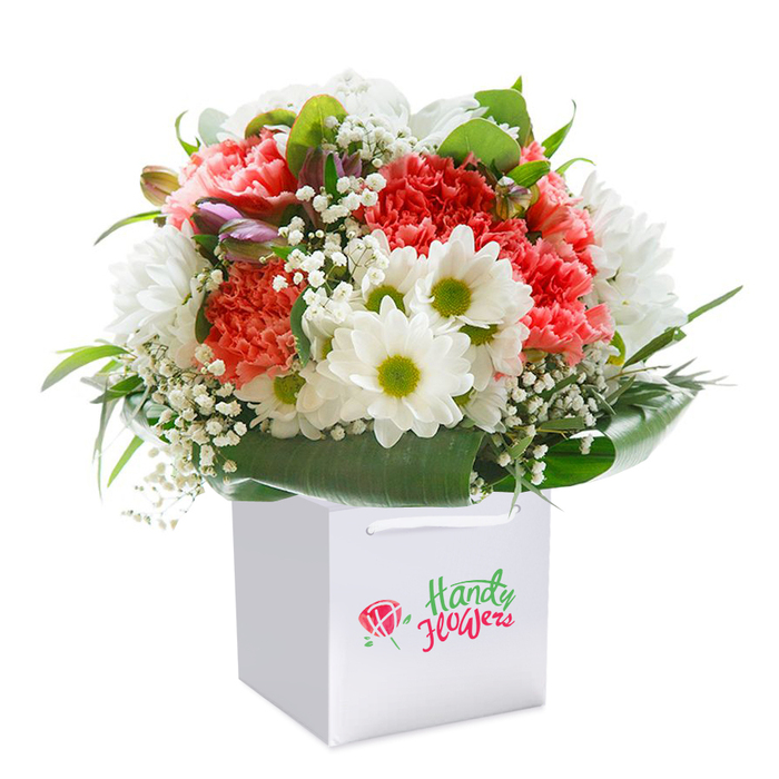 Expertly arranged in a sleek white gift bag, this vibrant Uxbridge bouquet celebrates abundance and artistry with its showcase of ruffled coral, peach, and crimson carnations layered beside crisp white daisies that catch the eye with their fresh yellow-green centers. Each carnation's frilly texture invites a closer look, while the gentle snowy petals of the daisies bring in a breezy, comforting calm-a nod to the gentle charm of Uxbridge's tree-lined streets. Wisps of delicate baby's breath thread through the arrangement, imparting subtle clouds of white that lighten and lift the overall form. Here and there, the arrangement reveals partially closed buds tinged in purplish-pink, adding a sense of anticipation and enhancing depth much like the changing light along Rockingham Recreation Ground. Circled at the base by sweeping ribbons of glossy, rich green foliage, the bouquet achieves a composed, rounded profile, putting every bloom on display. The accompanying gift bag, sturdy with double white handles, offers a gift-ready touch fitting for any celebration or noteworthy day-a graduation along Oxford Road, a new home, or a birthday surprise delivered to a favourite neighbour. The prominent branding on the bag, thoughtfully not overstated, echoes the reliable quality and bespoke beauty local to Uxbridge Flowers. Captured against an immaculate white background, the colours and craftsmanship of this arrangement stand out vividly, making it a true testament to the freshness and expertise behind every local delivery.